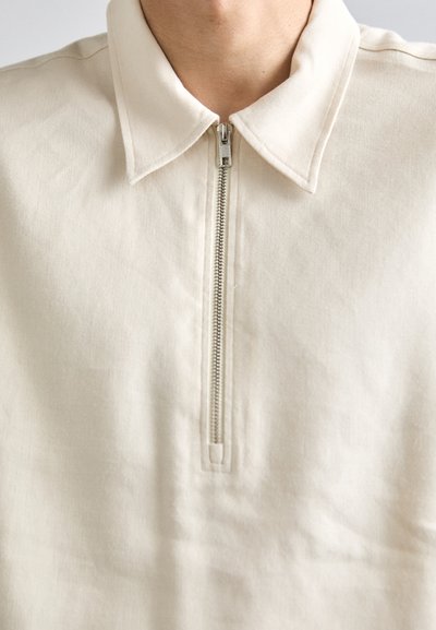 Pull-over beige à demi-fermeture éclair avec col, fabriqué à partir d'un tissu doux. Présente une fermeture éclair métallique et des coutures visibles le long des seams.