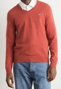 Homme portant un pull V en corail sur une chemise collier blanche, associé à un jean bleu, sur un fond uni.