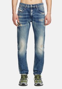 Jean délavé bleu clair à coupe droite, avec des déchirures sur les jambes et un léger effacement. Associé à des chaussures de sport grises et vertes.