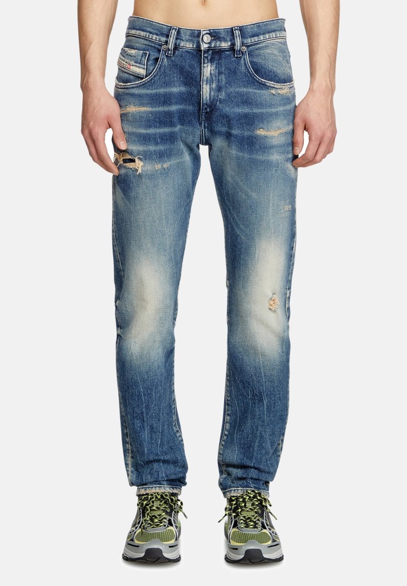 Jean délavé bleu clair à coupe droite, avec des déchirures sur les jambes et un léger effacement. Associé à des chaussures de sport grises et vertes.