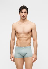 Νεαρός άνδρας με σκούρα σγουρά μαλλιά, όρθιος χωρίς μπλούζα, φοράει ανοιχτό γκρι στενά μπόξερ, μπροστά σε απλό φωτεινό φόντο.