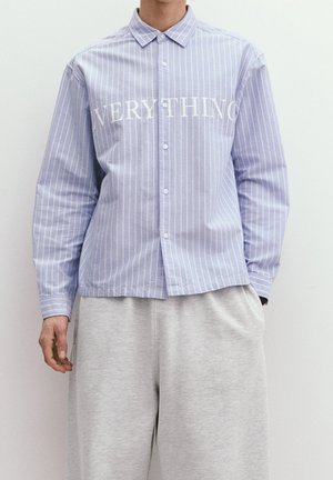 Chemise à rayures bleues et blanches à boutons avec le texte "EVERYTHING" en blanc sur la poitrine ; associée à un pantalon de survêtement gris.