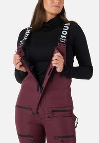 Burgundy skidragter med justerbare stropper, lynlåsede lommer og vandtæt materiale. Har en sort inderside og knapper til lukning.