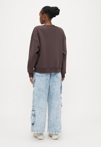 Brūns sasiets svīteris ar nolaistām pleciem, kopā ar gaiši ziliem plašiem cargo džinsiem ar vairākiem kabatām un nodilušu apdari.