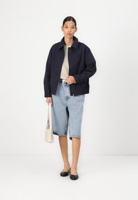 Jachetă de tip zip-up de culoare bleumarin, cămașă bej, șorturi din denim de culoare albastru deschis, pantofi joși negri și o mică geantă bej; material neted, croială lejeră, design minimal.