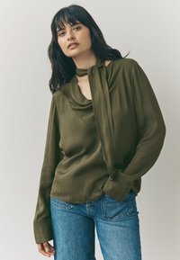 Olive green blouse with a draped neckline and tie detail. Long sleeves with wide cuffs, paired with light blue jeans. Smooth, silky texture.