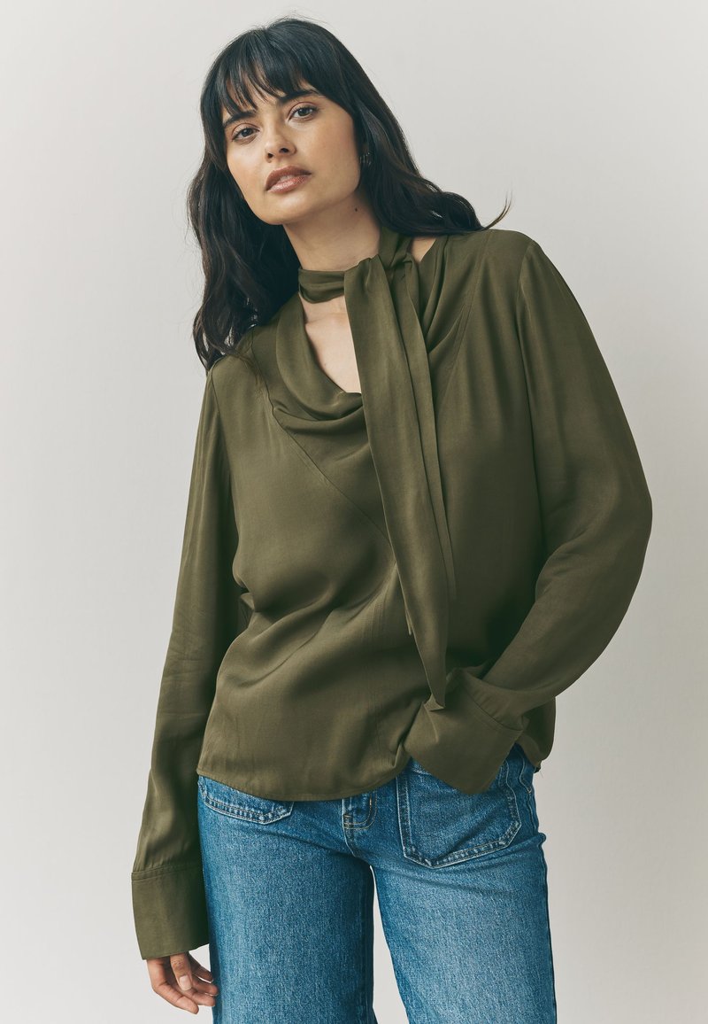 Olive green blouse with a draped neckline and tie detail. Long sleeves with wide cuffs, paired with light blue jeans. Smooth, silky texture.
