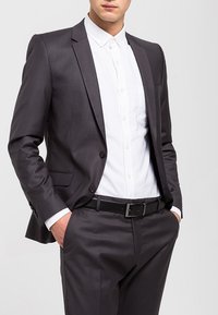 Chaqueta de traje gris oscuro con solapas en muesca y dos botones, combinada con una camisa blanca y un cinturón negro. Tela suave y corte entallado.