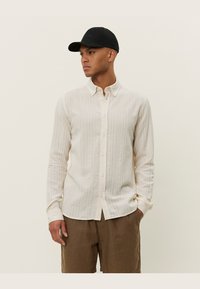 Lange mouw crèmekleurig shirt met een gestructureerd patroon, knoopsluiting aan de voorkant en een puntige kraag, gecombineerd met beige shorts en een zwarte pet.