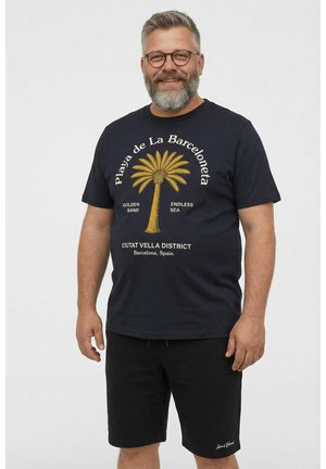 Uśmiechnięty mężczyzna w średnim wieku z okularami i siwą brodą, ubrany w czarną koszulkę z palemką i czarne szorty na jednolitym tle.