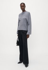 Grå oversized genser i myk strikk, kombinert med skreddersydde svarte bukser. Modellen holder en liten svart håndveske og har på seg spisse svarte sko.
