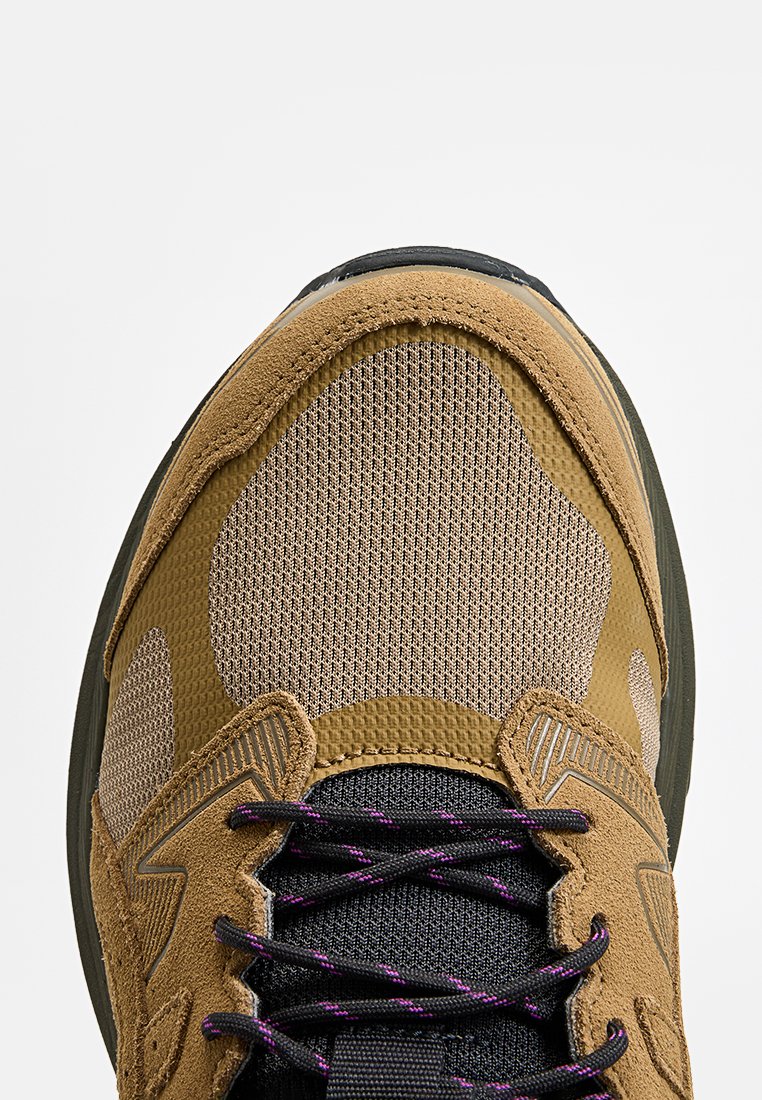 Gros plan sur l'embout d'une sneaker marron et beige avec des lacets noirs et violets, fabriquée en mesh et en daim, avec des coutures détaillées.