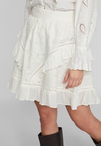 Bílé vyšívané sukni s květinovými výřezy a volánkovými okraji, kterou doplňuje odpovídající halenka s dlouhým rukávem a tmavohnědé kozačky až po kolena.