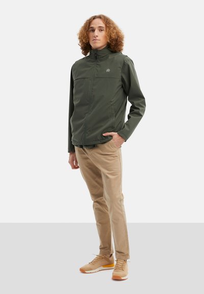 Zelená lehká bunda s vysokým límcem a předním zipem, béžové kalhoty a béžové tenisky, na hrudi bundy je viditelné logo.