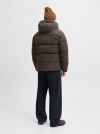 Rjava puhovka s kapuco, z vodoravnimi šivi, v kombinaciji z ohlapnimi črnimi hlačami in rebrastim rjavim klobukom.