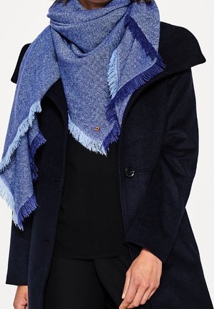 Personne portant un manteau bleu marine foncé et une grande écharpe bleue à franges enroulée autour du cou et des épaules.