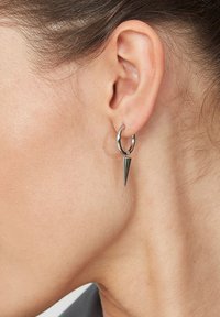 Sudrabaini auskari ar norādītu piekariņu, kas ir ar pulētu apdari un gludu tekstūru. Vienkārša, moderna dizaina pieeja, kas koncentrējas uz ģeometriskām formām.