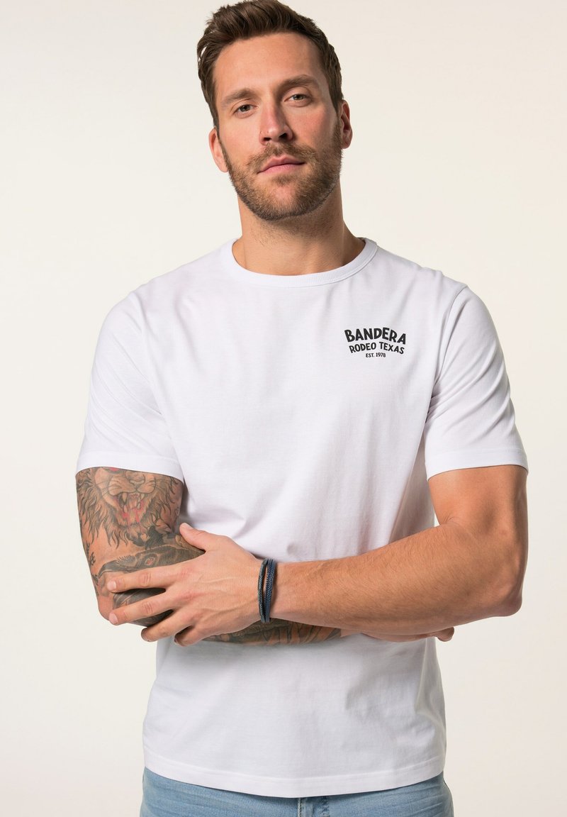 Camiseta de algodón blanca con cuello redondo, que presenta un logotipo impreso en negro "BANDERA RODEO TEXAS" en el pecho. Modelo con tatuajes visibles y pulsera.