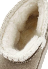 Bota de piel de oveja beige con forro de piel de crema, exterior de ante texturizado y borde de punto. Abertura superior redondeada, interior suave.
