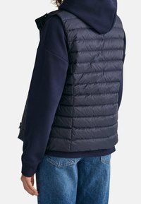 Marinblå puffväst med quiltad textur, med sidofickor och hög krage, bärs över en matchande marinblå hoodie och blåjeans.