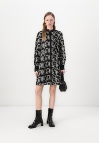 Vestido preto com bordado floral branco, gola alta e mangas longas. Combinado com botas pretas de salto e uma bolsa preta.