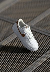 Bijela Nike Air Force 1 tenisica s smešnim swooshom i naglascima, prikazana na nagnutoj metalnoj rešetkatoj površini na suncu.