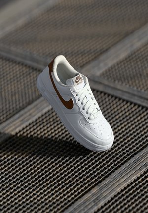 Bijela Nike Air Force 1 tenisica s smešnim swooshom i naglascima, prikazana na nagnutoj metalnoj rešetkatoj površini na suncu.
