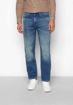 Man wearing blue faded jeans, brown checkered shirt, and white sneakers, standing against a plain light background.