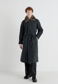 Un trench coat lung negru cu guler de blană, talie cu brâu și închidere cu nasturi. Țesătura are o textură netedă, asociată cu pantaloni și încălțăminte întunecată.