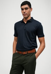 Camisa polo de color azul marino con cuello y mangas cortas, combinada con pantalones verde oliva y un cinturón de cuero marrón. Textura de tela suave.