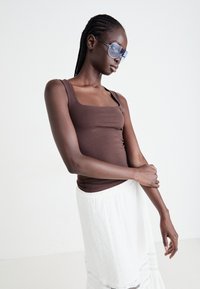 Brun genser med firkantet utringning, kombinert med et hvitt brodert skjørt. Modellen bruker oversized blå solbriller.