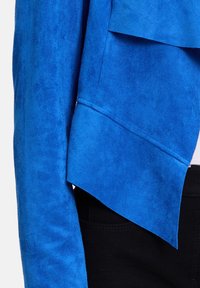 Gros plan de la manche d'une veste en daim bleu et détail de rabat superposé sur un pantalon noir.