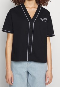 Camicia nera a maniche corte con scollatura a V, caratterizzata da una bordatura bianca e una chiusura con bottoni frontale. Dettaglio ricamato "Tommy 85" sul petto.