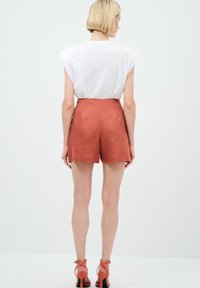 Pantaloni corti in lino ruggine con vita alta e tasche laterali; abbinati a una maglietta bianca. Modella con tacchi a strappo, vista da dietro.