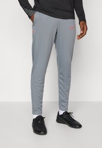 Gray athletic pants with a slim fit, featuring an orange Nike logo and zippered pockets; made of smooth, lightweight fabric.