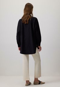 Una donna con capelli di lunghezza spalla sta in piedi di spalle indossando una maglietta nera larga, pantaloni cropped crema e ballerine beige con punta.