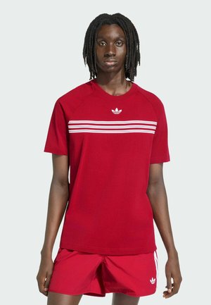 Młoda osoba o ciemnej karnacji i dredach, ubrana w czerwoną koszulkę Adidas z białymi paskami oraz pasujące czerwone szorty z logo Adidas.