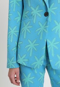 Veste bleu clair avec motif de palmier vert, fermeture à un bouton et détail de poche avant. Le tissu texturé ajoute un intérêt visuel.