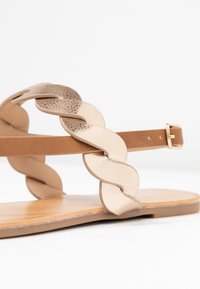 Flade sandaler med en glat tan sål, der har en flettet rem i lyse beige med en struktureret overflade og en sidelukning til justering.