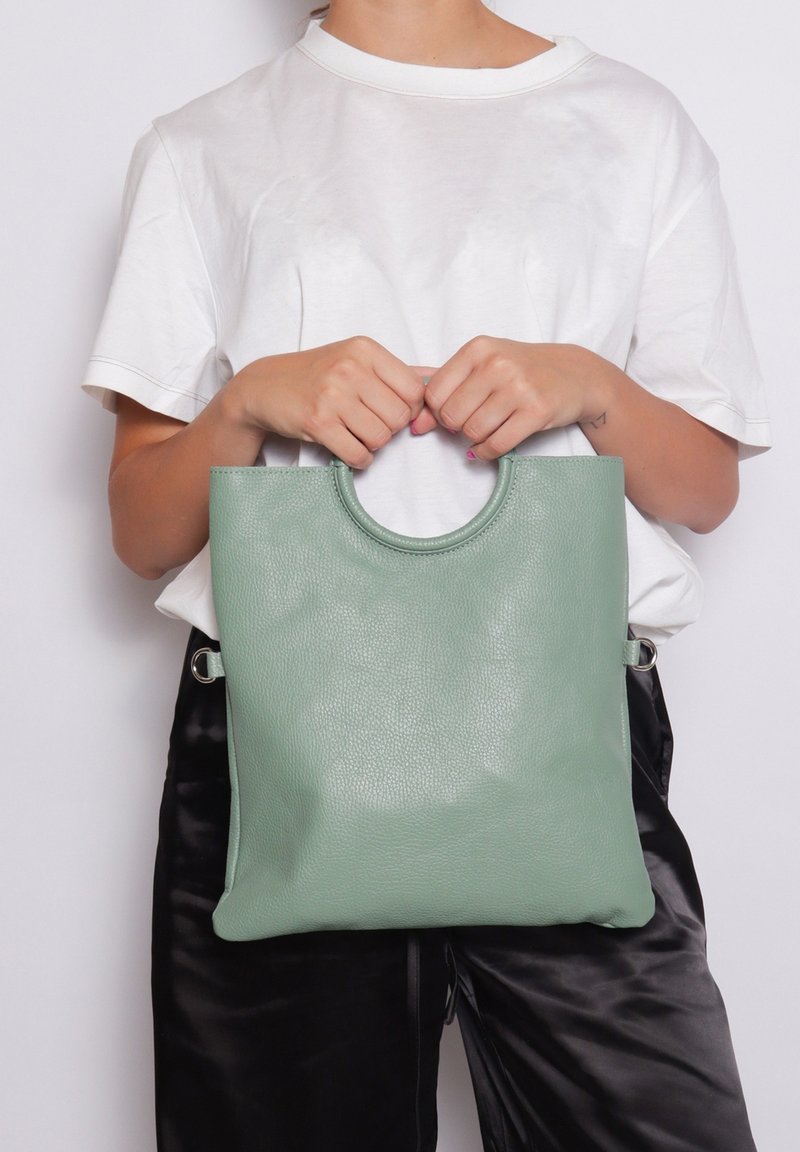 Borsa a tracolla verde in pelle con manico arrotondato, texture liscia e dettagli a cerchio sui lati, tenuta da una persona in abbigliamento informale.