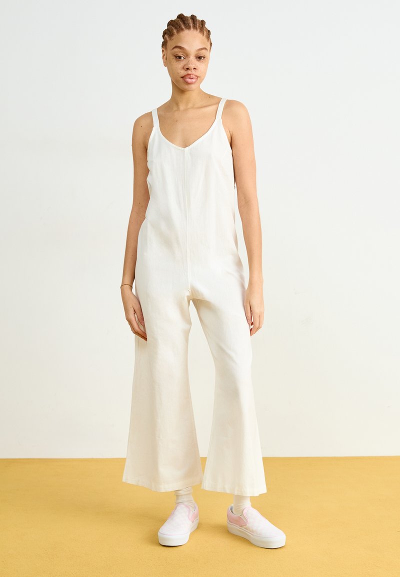 Mono de lino blanco con escote en V, tirantes finos y diseño de pierna ancha. La modelo lleva zapatillas blancas con toques de rosa y calcetines hasta el tobillo.