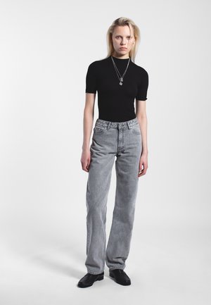 Schwarzes kurzärmeliges Oberteil, kombiniert mit hoch taillierten, hellgrauen Jeans. Das Model trägt mehrere silberne Ketten und schwarze, flache Schuhe.