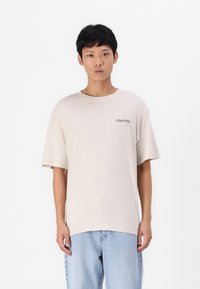 Beige kortermet T-skjorte med rund hals og et trykt "Club1990"-logo på venstre side. Kombinert med lyseblå denimjeans.