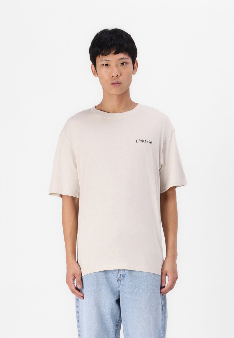 Beige kortermet T-skjorte med rund hals og et trykt "Club1990"-logo på venstre side. Kombinert med lyseblå denimjeans.