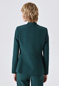 Blazer su misura verde scuro con collo alto, spalle strutturate e fondo leggermente svasato. Tessuto liscio con dettagli di cucitura puliti.