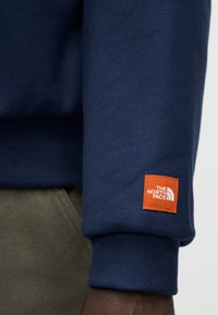 Ναυτικό μπλε φούτερ με εξάρτημα σε πορτοκαλί τετράγωνο και το λογότυπο "The North Face" στο μανίκι. Μαλακό ύφασμα με λεπτομέρεια ριμπ στο μανσέτι.