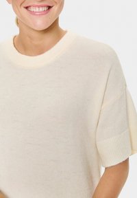 Personne souriante portant un pull en tricot à manches courtes de couleur crème, avec un col et des poignets côtelés, sur fond blanc uni.