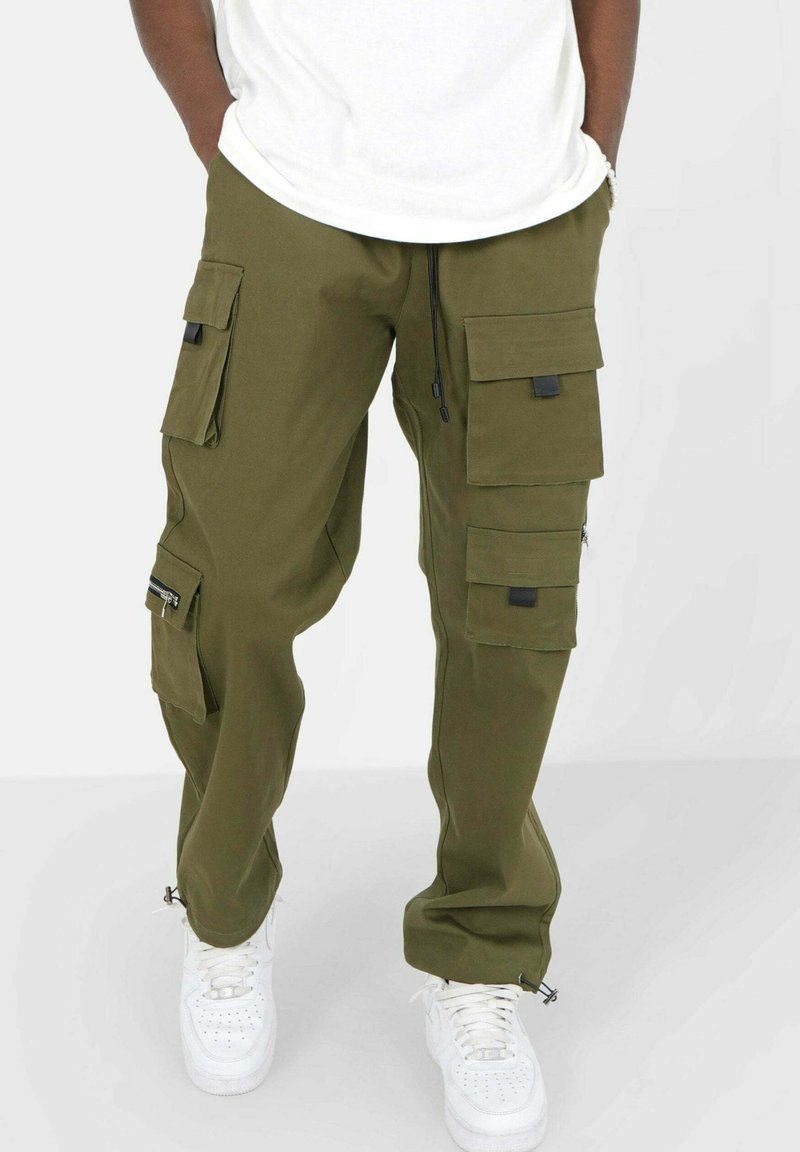 Olijfgroene cargobroek met meerdere klepzakken, verstelbare koord in de taille en ritsen bij de enkels, gecombineerd met witte sneakers.