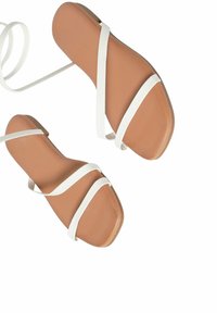 Sandali con cinturini, con una soletta color bronzo e cinturini bianchi. La suola è piatta, con una texture liscia e una punta rotonda.