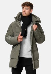 Puffer jacket in olive green with a quilted design, featuring a hood and pockets. Worn over a light gray sweater and black scarf.